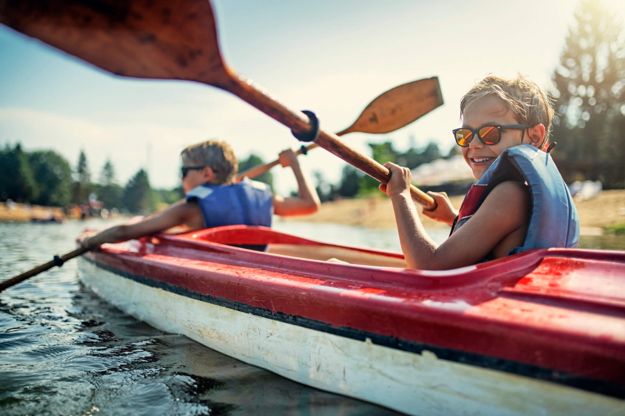 Two children sitting in a canoe on water, holding oars and wearing sunglasses and life jackets.